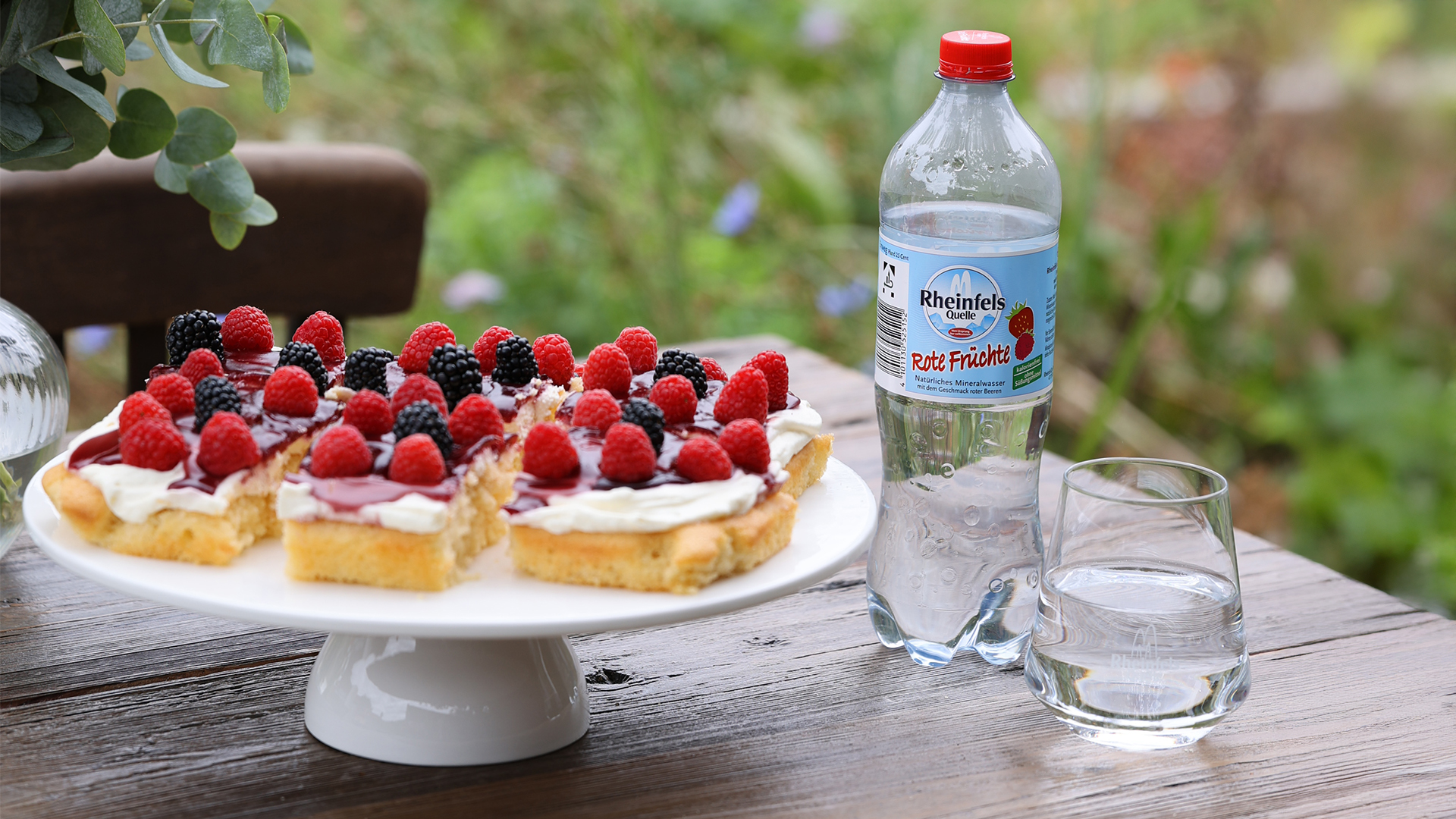 Mineralwasser-Kuchen mit Beeren-Mix auf einem Tortenboden angerichtet, dazu auf dem Tisch stehend und eingeschenkt in ein Glas, Rheinfels Quelle Rote Früchte in einer Einweg Flasche.