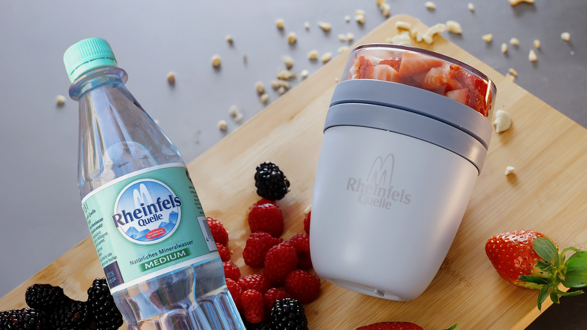 Overnight Oats im Lunchpot to go mit einer Flasche Rheinfels Quelle Medium 0,5 Liter PET Mehrweg. Beides auf einem Holzschneidebrett dekoriert mit Früchten.