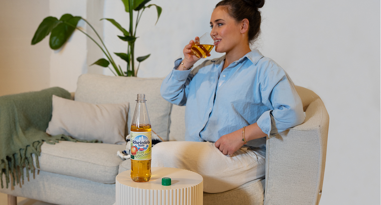 Eine Frau sitzt auf der Couch und genießt aus einem Glas ihre Erfrischung - Milde Apfelschorle.