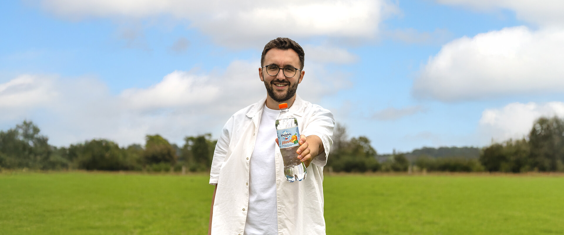 Ein junger Mann hält eine Flasche Rheinfels Quelle Pfirsich in die Kamera, im Hintergrund ist eine Wiese zu sehen.