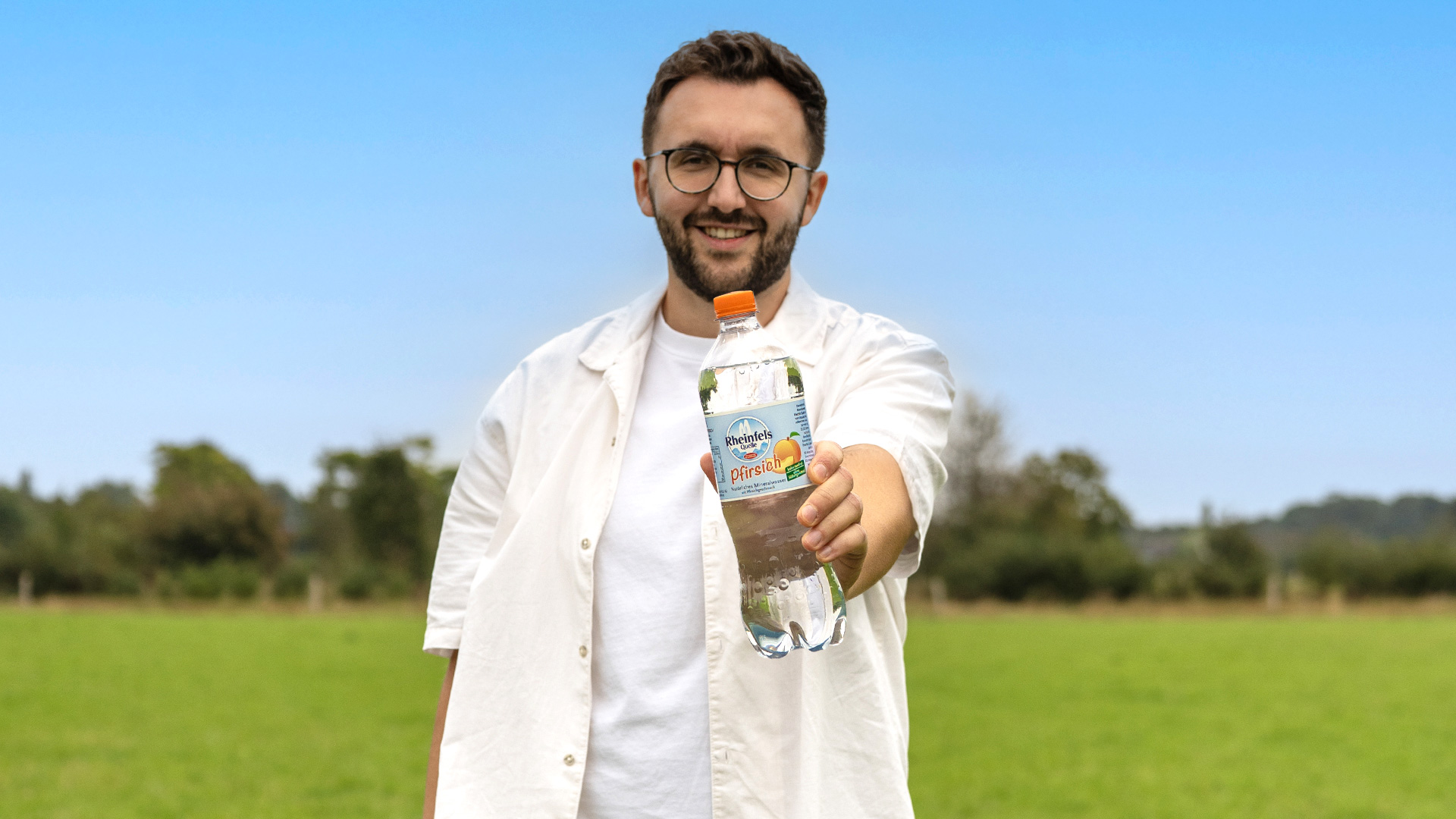 Ein junger Mann hält eine Flasche Rheinfels Quelle Pfirsich in die Kamera, im Hintergrund ist eine Wiese zu sehen.