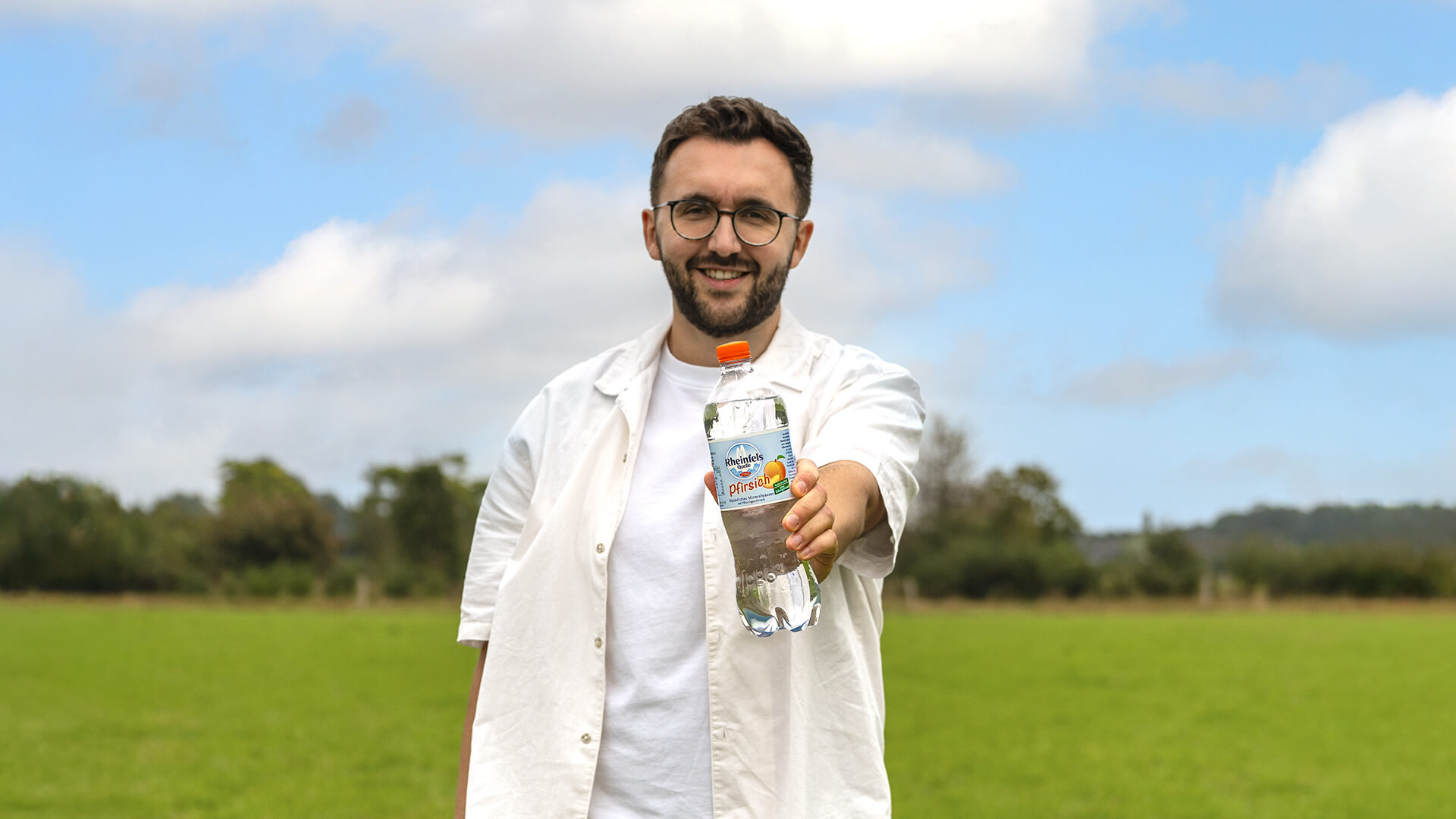 Ein junger Mann hält eine Flasche Rheinfels Quelle Pfirsich in die Kamera, im Hintergrund ist eine Wiese zu sehen.