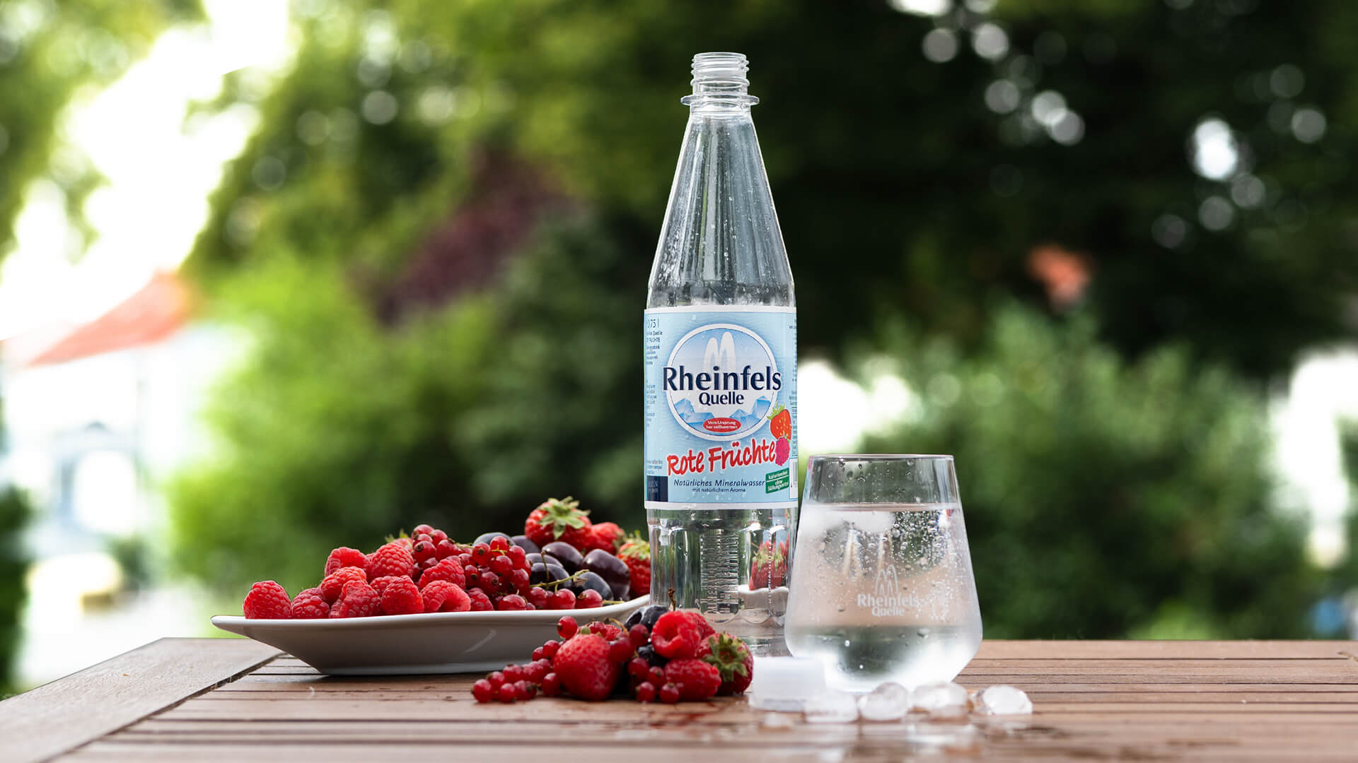 Auf einem Balkon steht ein Tisch mit einer Flasche Rote Früchte umgeben von Früchten und Eiswürfeln. Davor steht ein gefülltes Glas mit dem aromatierstem Mineralwasser Rote Früchte.