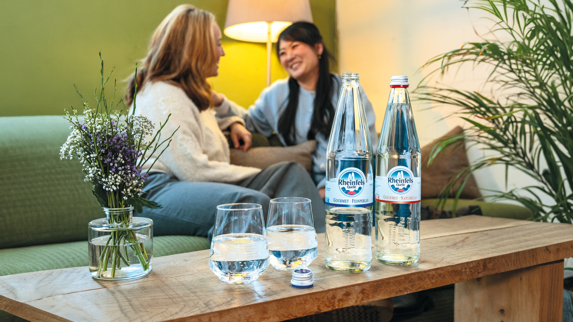 Zwei Frauen auf einem Sofa, im Vordergrund zwei gefüllte Gläser mit Rheinfels Quelle Gastronomie Feinperlig und Naturelle Flaschen auf dem Tisch