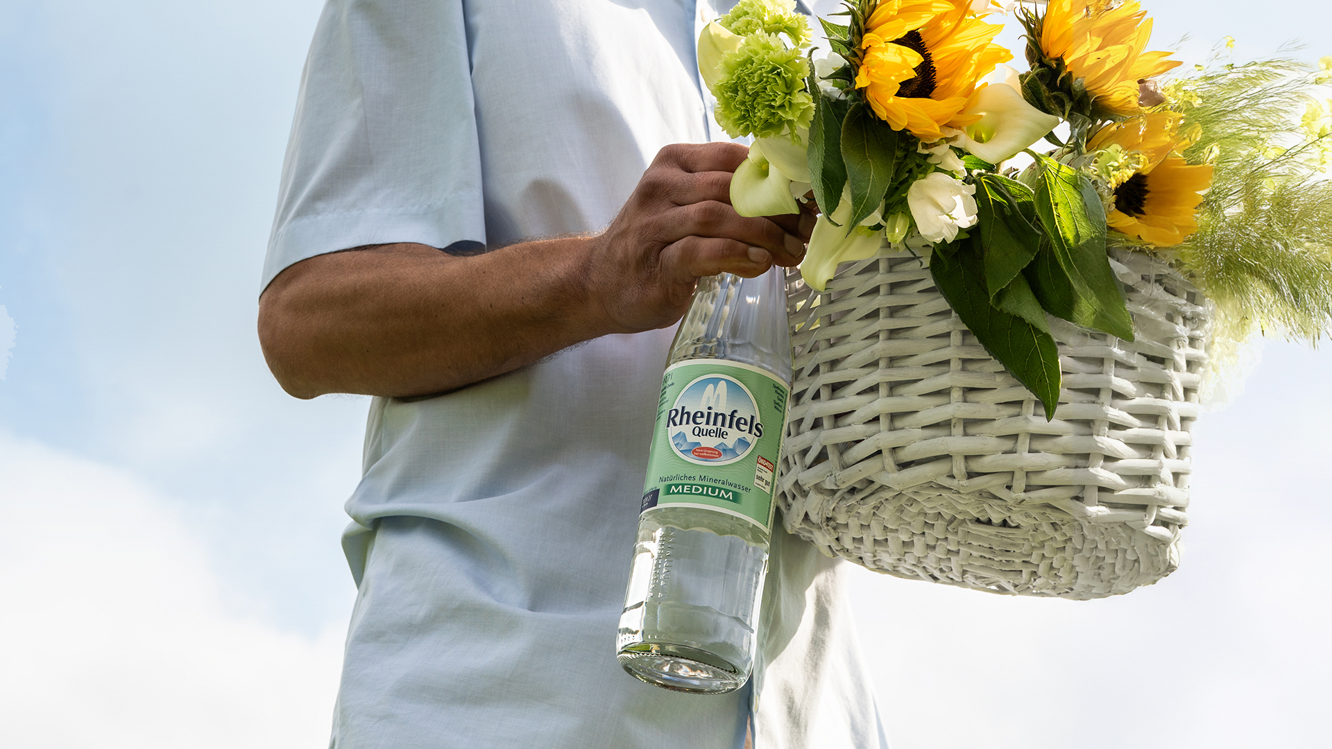 Ein Mann ist auf dem Weg zu einem Dinner. Er hat einen weißen Weidenkorb dabei und hält eine Rheinfels Quelle Medium Glasflasche in der Hand