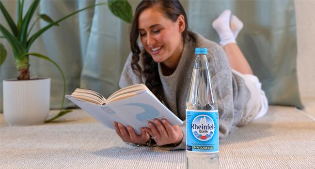 Eine Frau liegt in ihrem Wohnzimmer auf dem Fußboden und liest ein Buch. Eine Flasche Rheinfels Quelle Naturelle 0,7 Liter Glas steht auf dem Teppich.