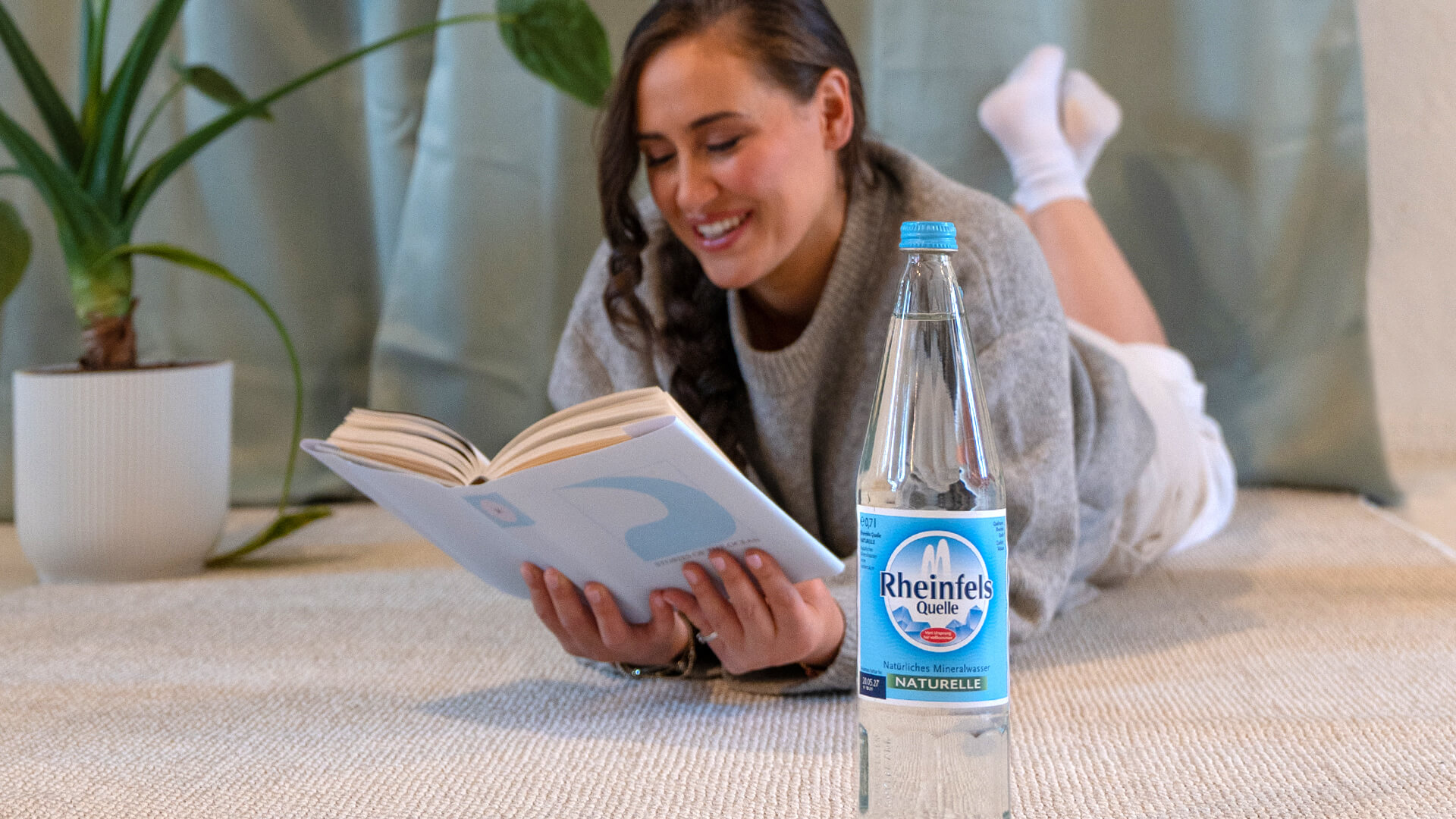 Eine Frau liegt auf einem Teppich und liest, eine Glasflasche Rheinfels Quelle Naturelle steht neben ihr.