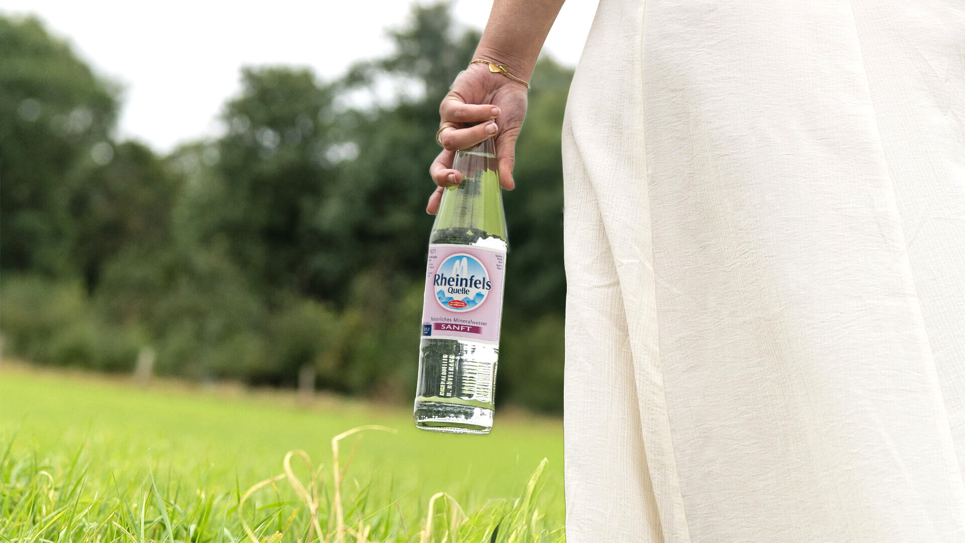 Eine Frau hält eine Rheinfels Quelle Sanft Glasflasche in der Hand, im Hintergrund ist Natur zu sehen